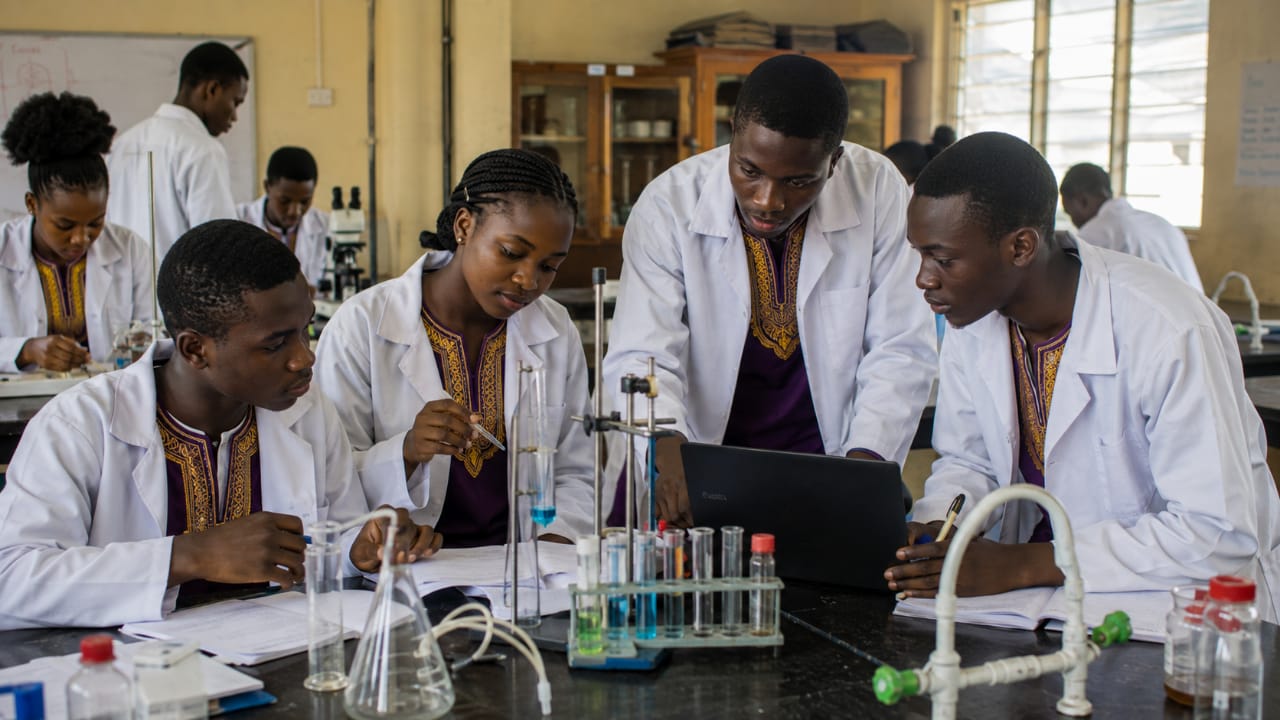 Nigerian secondary school learners in lab coats over dashiki outfits collaborating on a practical science investigation using laboratory equipment, notebooks, and a computer.